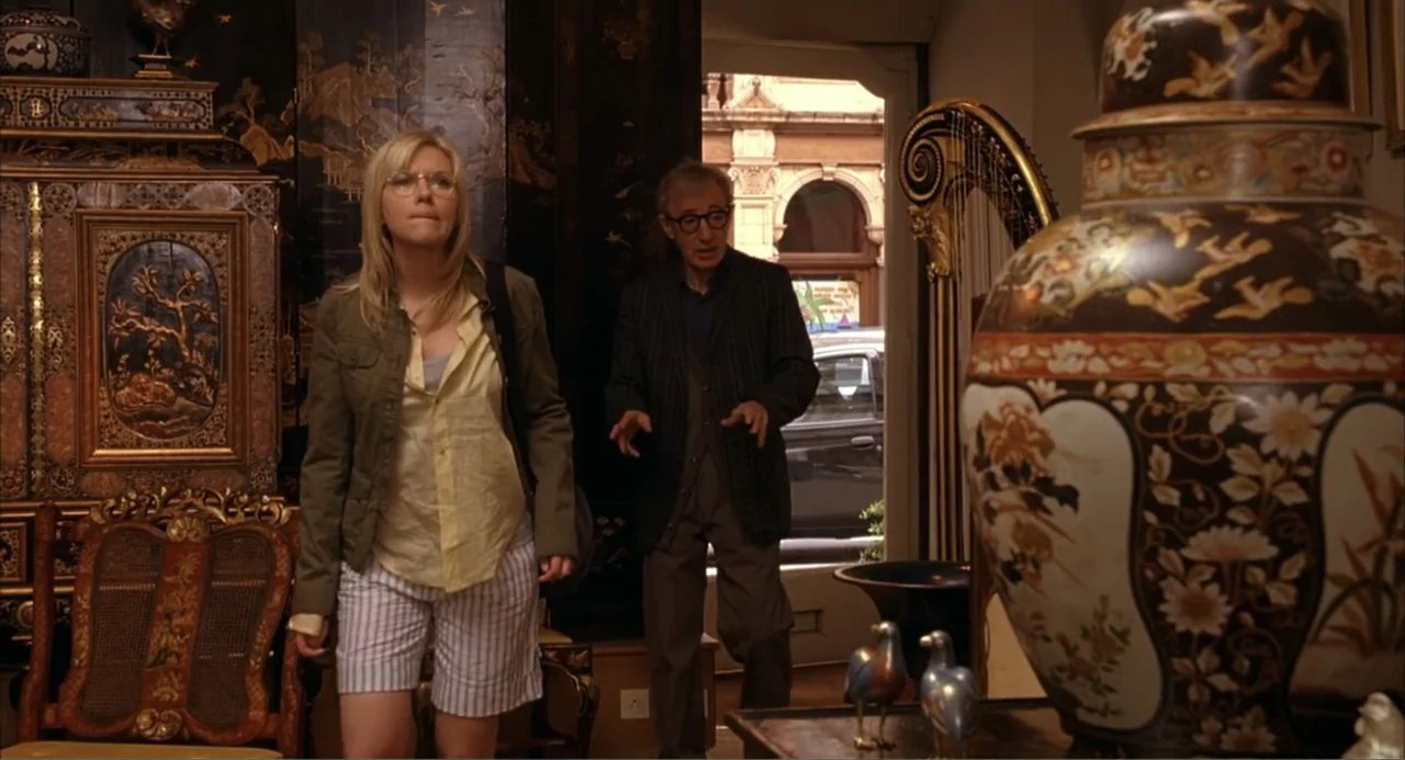 Scoop (2024) movie still: medium — An interior antique shop filled with ornate wooden furniture, large ceramic vases, and screens. A young woman and…