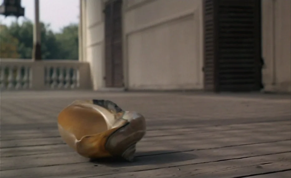 The Immortal Story (1968) movie still: closeup — A large seashell sits on the wooden planks of a wide, empty outdoor porch. White railings and…