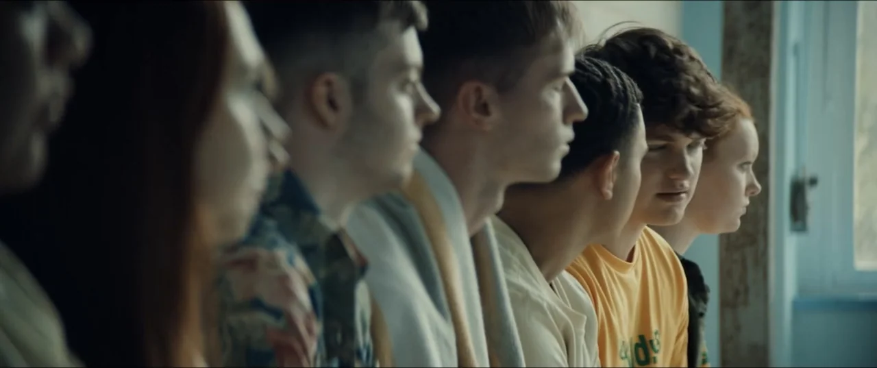 Bonus Track (2024) movie still: medium — A row of teenagers sits facing the same direction in a bright interior space. They are lined up…