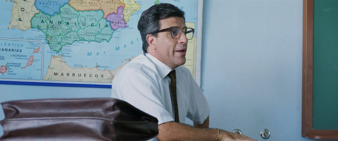 Voy a pasármelo bien (2022) movie still: medium — A classroom interior features a large colorful map pinned to a pale blue wall. A man in a…