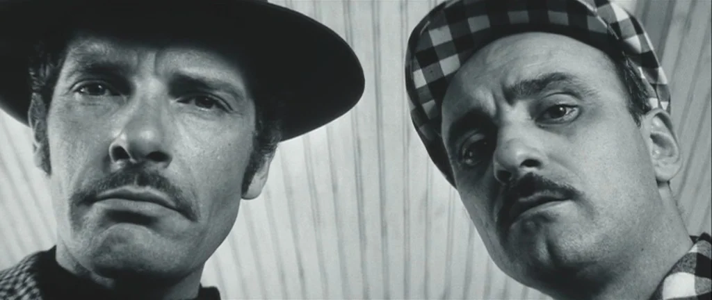 Les Créatures (1966) movie still: closeup — Two men stare downwards into the camera lens against a vertical wooden paneled surface. The composition…