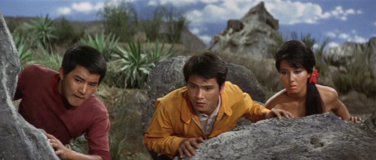 Ebirah Horror of the Deep (1966) movie still: medium — Three individuals crouch behind large grey boulders in a dry, sparse, rocky landscape. They…