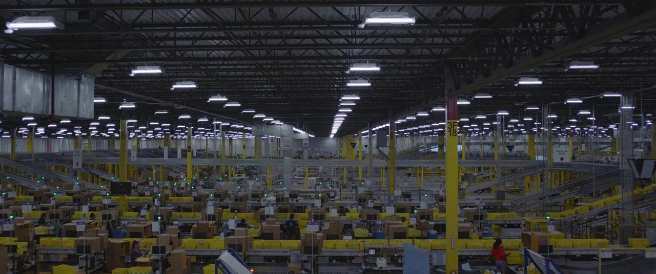 Nomadland (2020) movie still: establishing — A massive, sprawling industrial distribution warehouse features endless rows of yellow bins and…