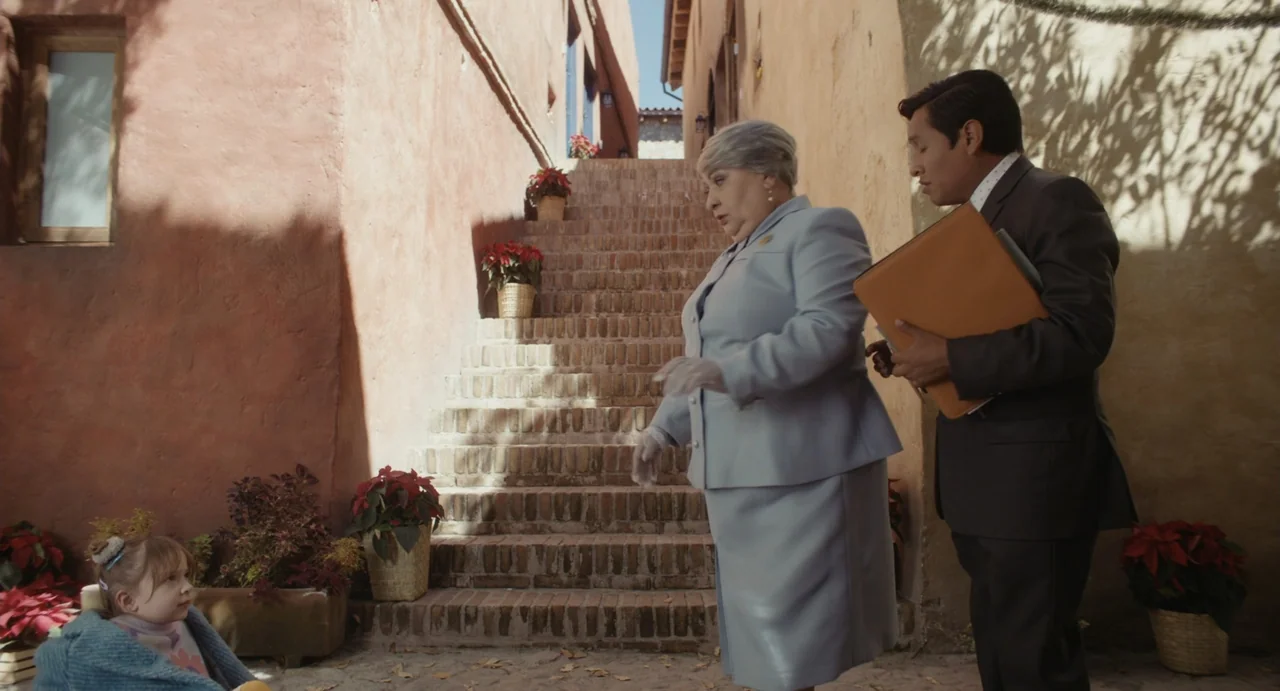 Dating Santa (2023) movie still: medium — A terracotta-walled alleyway features stone steps flanked by potted poinsettias under bright sunlight. An…