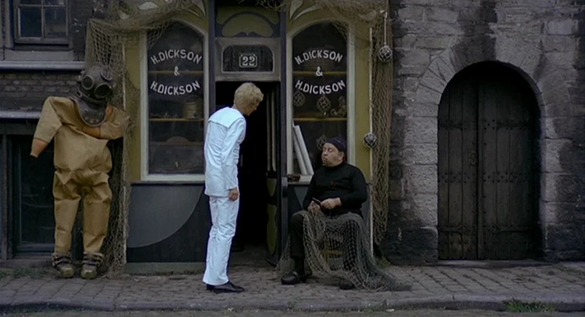 Malpertuis (1971) movie still: fullbody — A stone-walled building exterior features a shop entrance decorated with nautical fishing nets. A man in a…