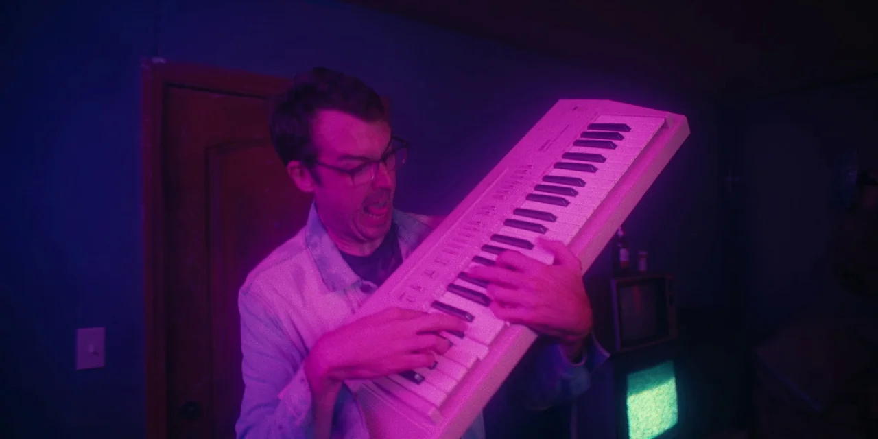 Destroy All Neighbours (2024) movie still: medium — A dimly lit room bathed in intense neon pink and blue light. A man holds a white synthesizer…
