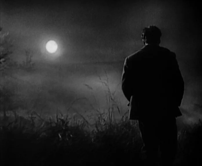 Sunrise (1927) movie still: medium — A dark, misty field at night is lit by a full, bright moon. A man in a dark suit stands with his back to the…