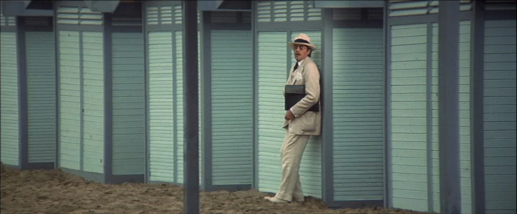 Death In Venice (1971) movie still: fullbody — A row of light blue wooden beach cabins stands on a sandy terrain. A man in a light-colored linen…