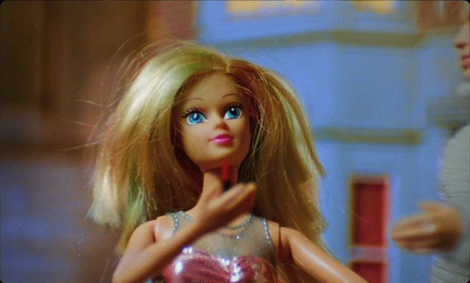 Misunderstood (2014) movie still: closeup — A doll with blonde hair stands inside a miniature structure with blue walls. The doll holds a small red…
