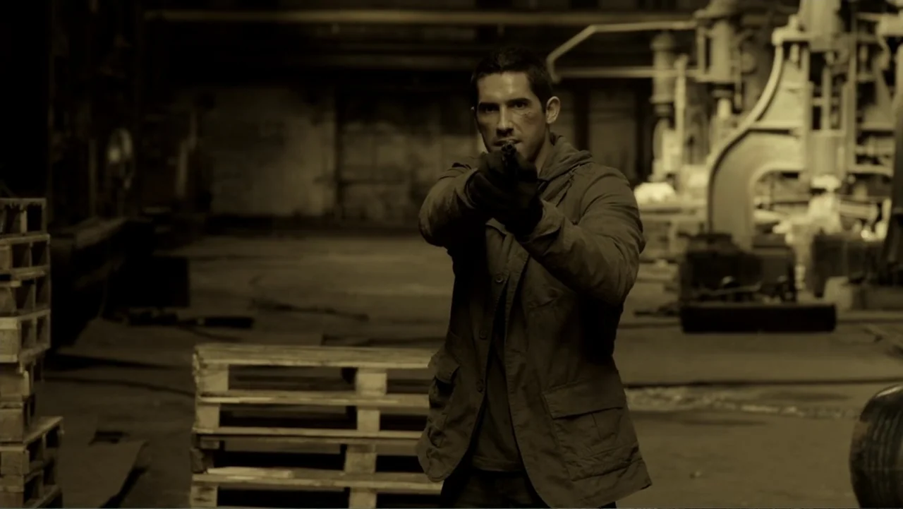 Assassination Games (2011) movie still: medium — A dark, industrial interior space filled with wooden pallets and heavy machinery silhouettes. A man…