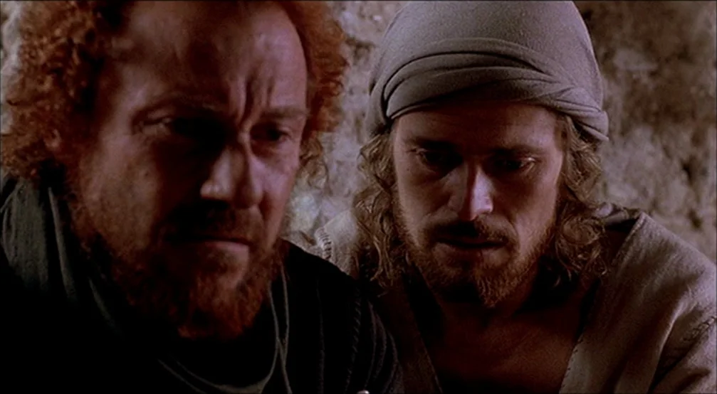 The Last Temptation Of Christ (1988) movie still: closeup — Two men sit close together in a dimly lit, rustic stone chamber. They both gaze…