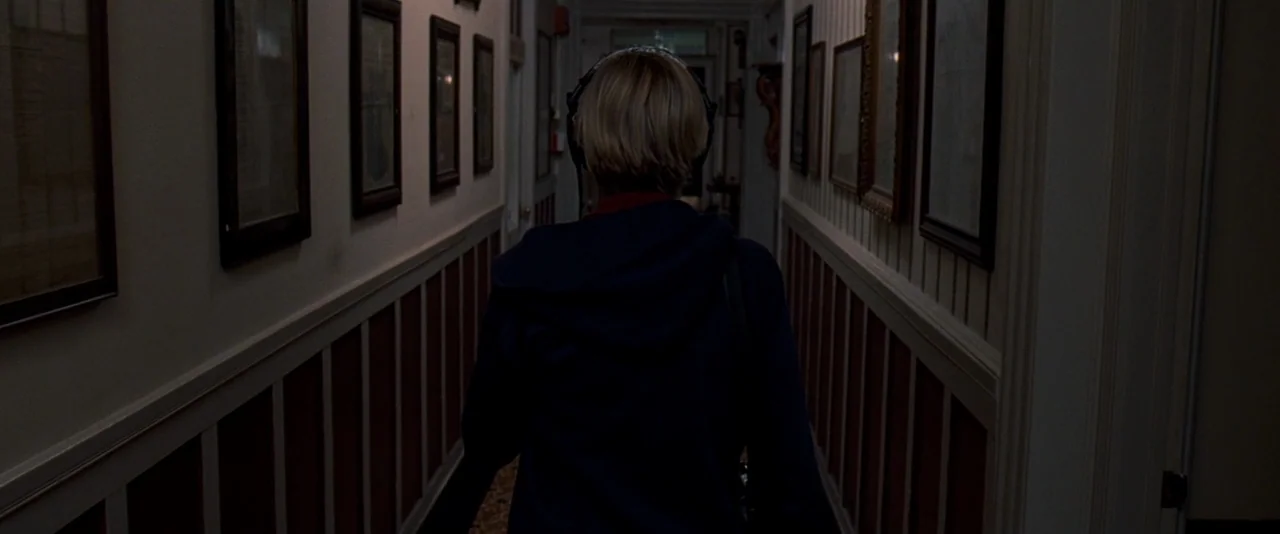 The Innkeepers (2011) movie still: medium — A narrow residential hallway features striped wallpaper and multiple framed pictures on both walls. A…