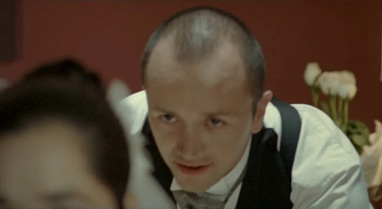 Sex, Love And Other Perversions II (2006) movie still: medium — A dimly lit dining area features a dark red wall and out-of-focus floral…