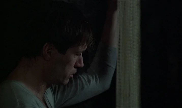 The Serpent’s Egg (1977) movie still: medium — A man stands in a dimly lit, nondescript interior space near a vertical textured frame. He leans his…