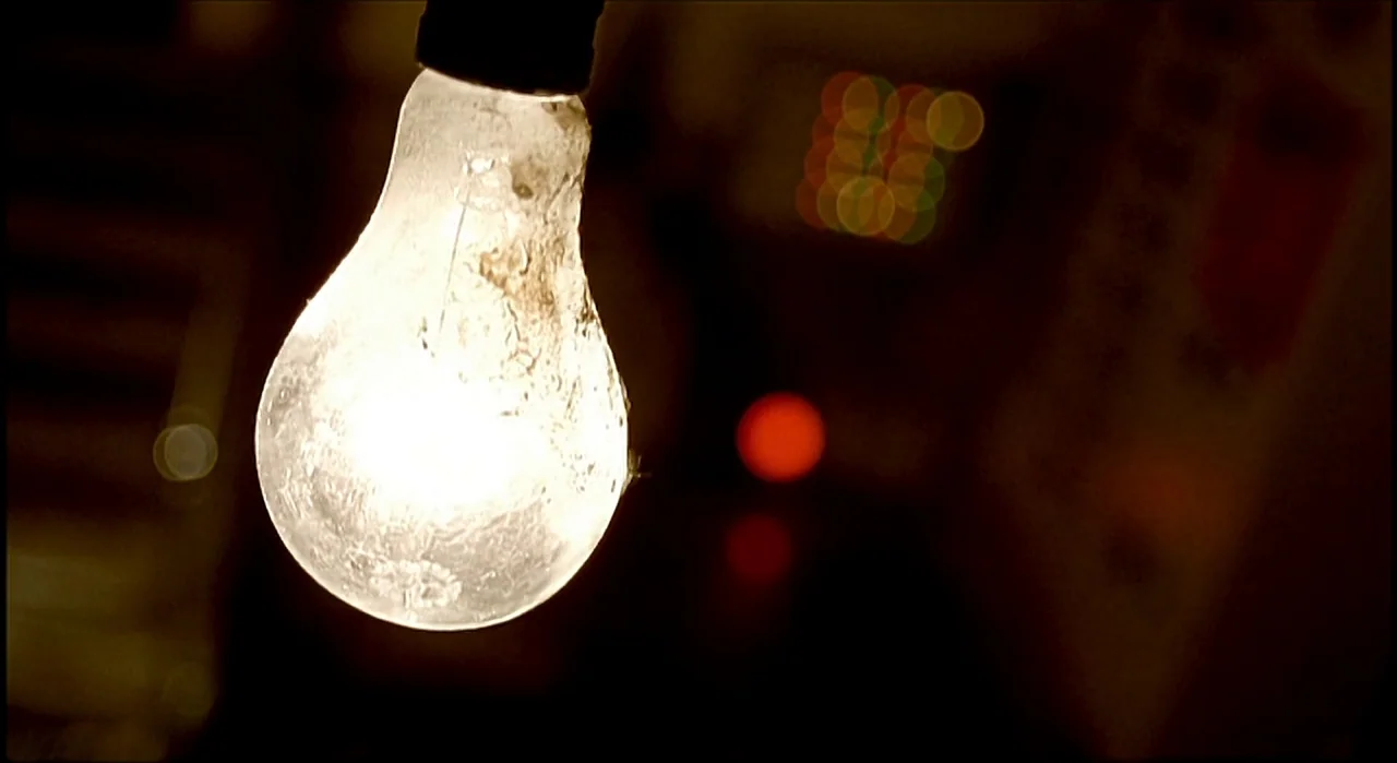 Voice (2005) movie still: closeup — A dusty incandescent light bulb hangs prominently in a dark, out-of-focus indoor space. The background features…