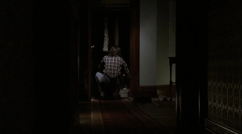 The House Of The Devil (2009) movie still: medium — A dimly lit hallway leads into a dark doorway where a person crouches. The floor is covered by a…