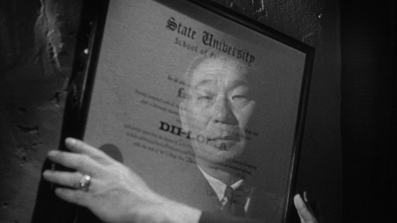 Shock Corridor (1963) movie still: closeup — A dark indoor space contains a framed university diploma held by unseen hands. The glass surface of the…