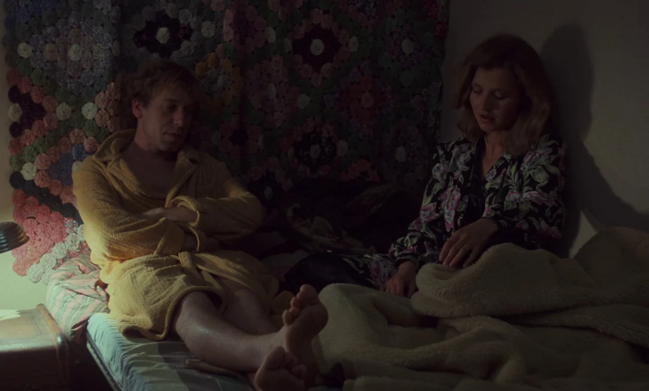 Wrong Move (1975) movie still: medium — A dark, dimly lit space features a bed with a colorful patchwork quilt wall hanging. A man in a yellow robe…
