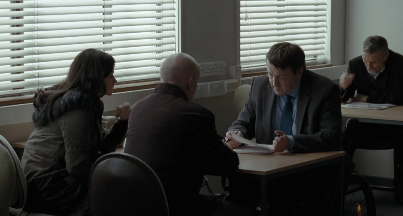 I, Daniel Blake (2016) movie still: medium — A functional room features wooden tables, horizontal blinds, and white electrical outlets on walls. A…
