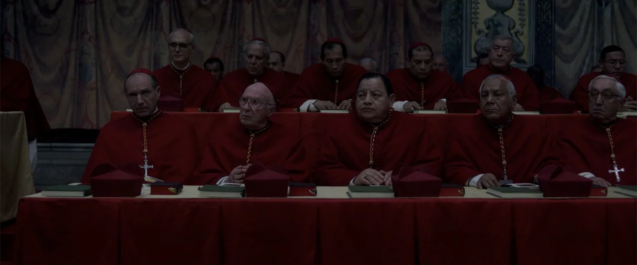 Conclave (2024) movie still: medium — A formal interior with red-clad cardinals seated behind a long table facing the camera. The individuals sit in…