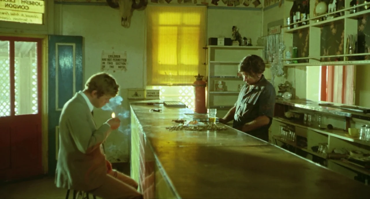 Wake In Fright (1971) movie still: medium — A rural hotel bar features wood counters, aged wall decor, and a yellow window blind. A man in a suit…