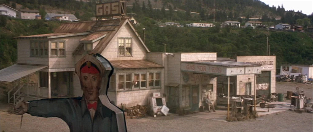 The Pledge (2001) movie still: wide — A weather-beaten rural gas station features white siding and a prominent roof sign. A two-dimensional…