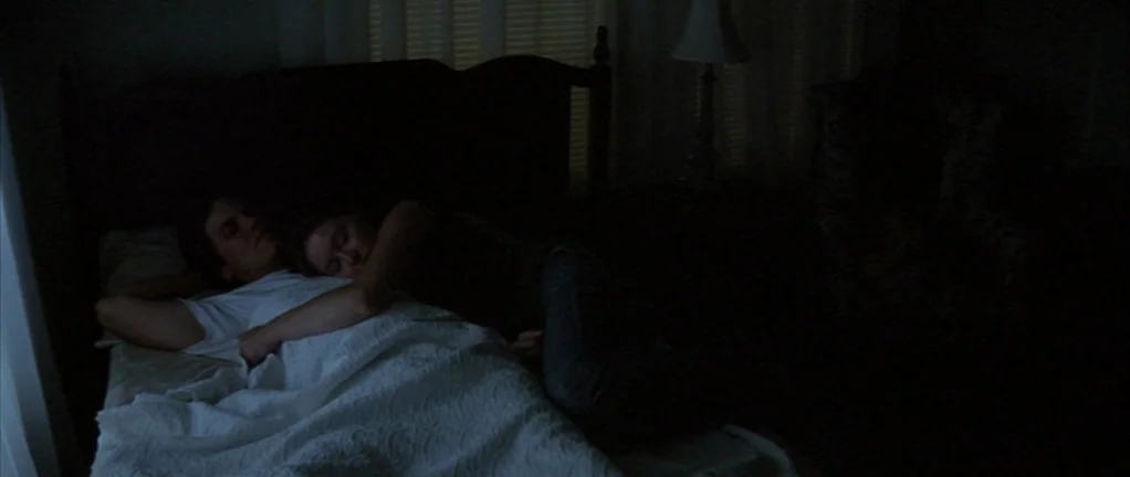 Shotgun Stories (2007) movie still: medium — A dark, minimally lit bedroom features a bed with white bedding and a lamp. A man and a woman lie…