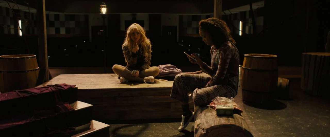 Freaky (2020) movie still: wide — A dimly lit theatrical stage features a wooden platform and a log prop. Two young women occupy the space, one sits…