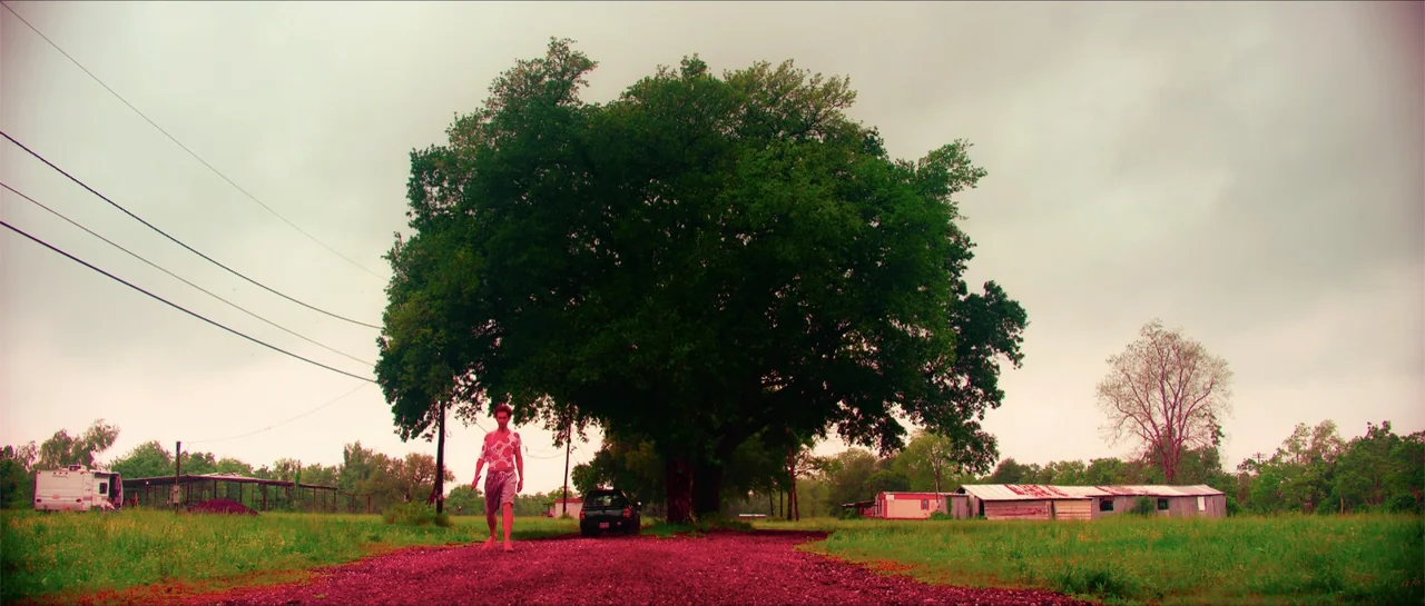 68 Kill (2017) movie still: wide — A rural expanse features a large central tree, a dirt road, and distant mobile homes. A solitary man in casual…