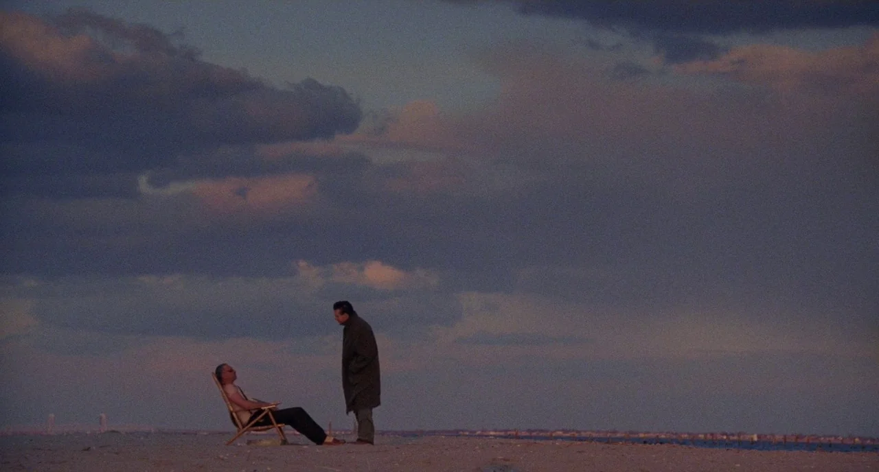 Angel Heart (1987) movie still: wide — A vast, empty beach during the dusk hours features large dark clouds above. Two men stand and sit on the sand…