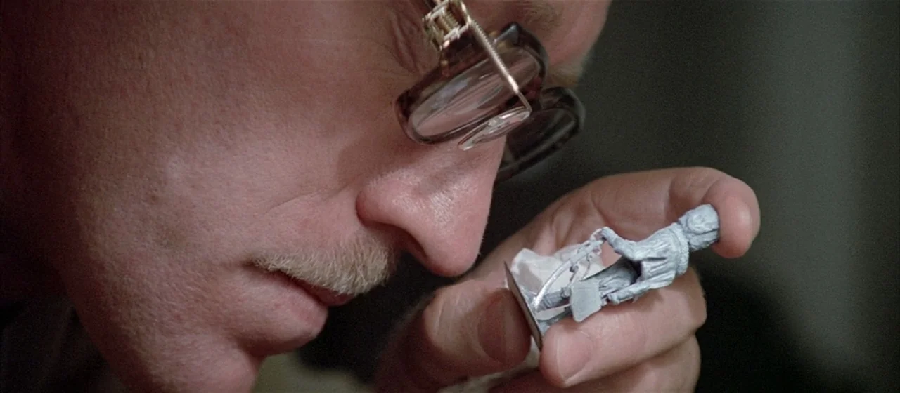 Three Days of the Condor (1975) movie still: closeup — A close-up view shows a person examining a small unpainted figurine closely. The man wears…