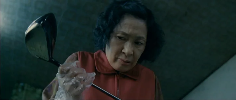 Mother (2009) movie still: closeup — A dimly lit room with muted wall patterns and shadows. An elderly woman grips a golf club while wearing a clear…
