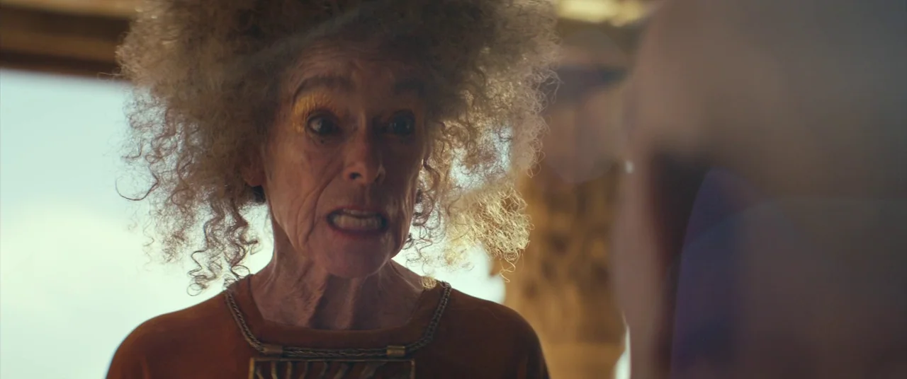Seneca: On The Creation Of Earthquakes (2023) movie still: closeup — A woman with frizzy hair stands in a brightly lit interior space. She stares…