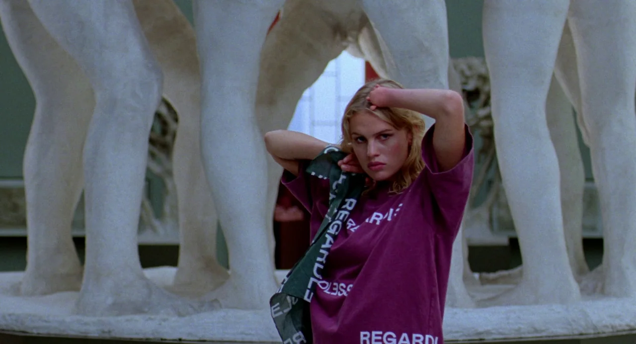 Sick of Myself (2022) movie still: medium — A woman stands in front of large, pale, classical-style statues in a public hall. She looks directly…