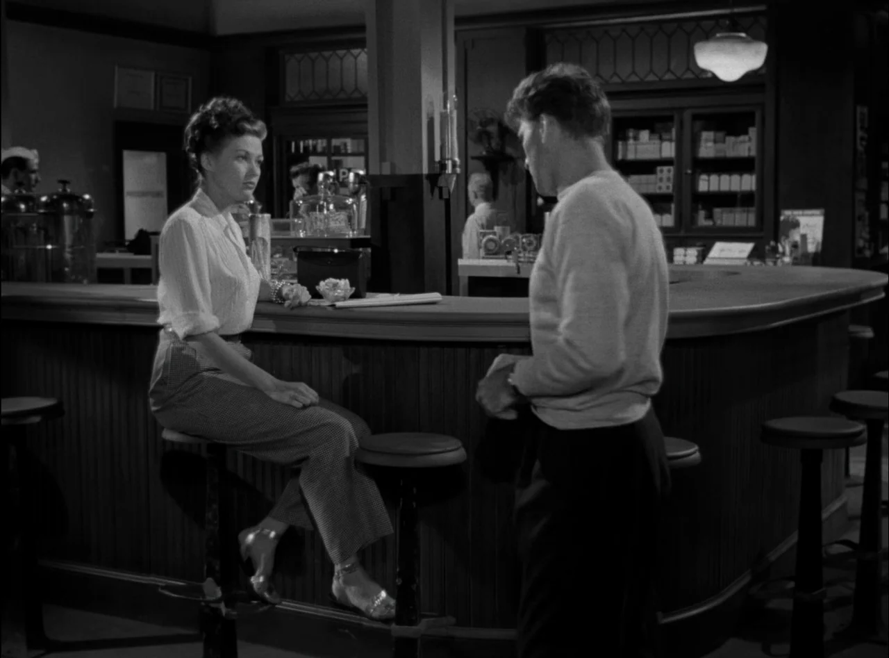 Criss Cross (1949) movie still: medium — A 1940s soda fountain features a curved wooden counter with round stools and shelving. A woman sits on a…