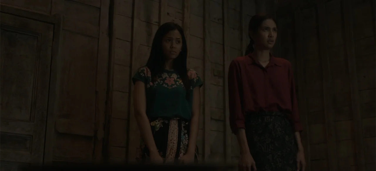 A Thousand Days (2023) movie still: medium — A dimly lit room features walls constructed from vertical planks of aged, dark wood. Two young women…