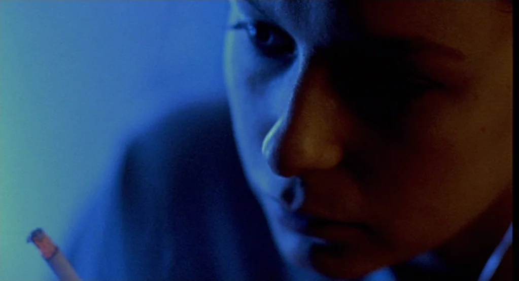 Morvern Callar (2002) movie still: closeup — A dimly lit interior space is saturated in a deep blue color cast. A young woman holds a cigarette…