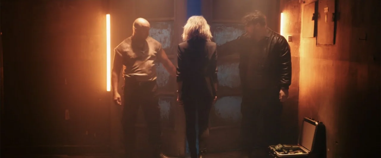 Jolt (2021) movie still: fullbody — An dimly lit industrial space with textured walls and vertical light fixtures. Three people stand together…
