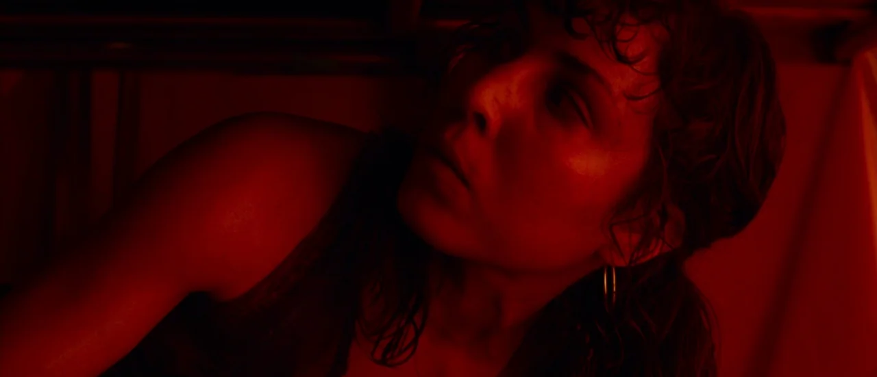 Rupture (2016) movie still: closeup — A dimly lit interior space is bathed in intense red light. A young woman with damp hair gazes upward towards…