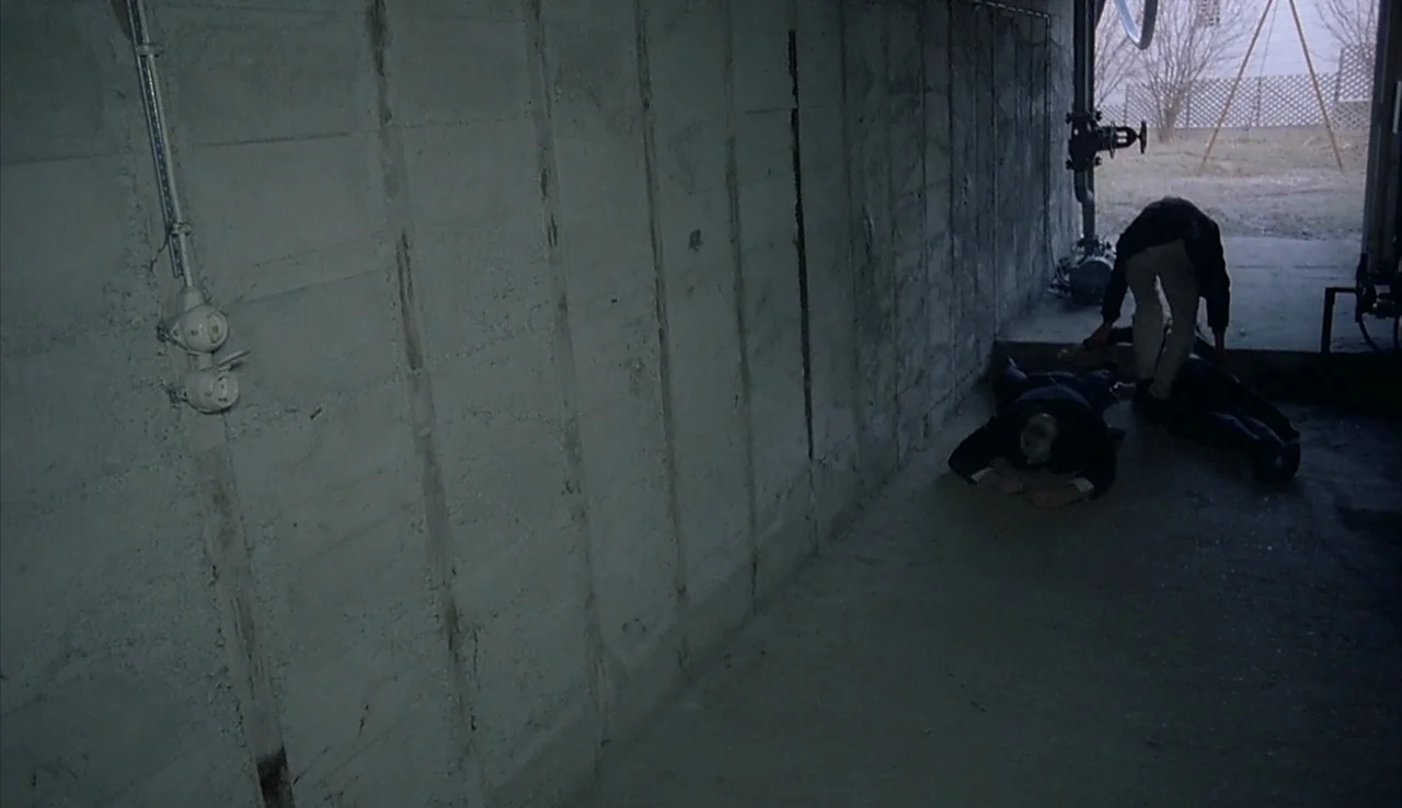 Angst (1983) movie still: wide — A bare concrete subterranean space features heavy industrial pipes near an open exterior door. One person stands…