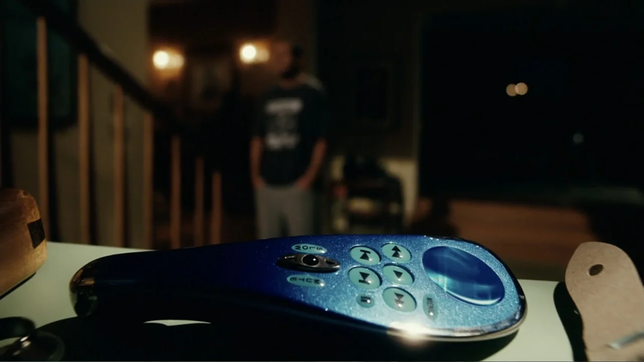 Click (2006) movie still: closeup — A dark indoor setting features a blurred figure standing in the background. A blue remote control lies on a…