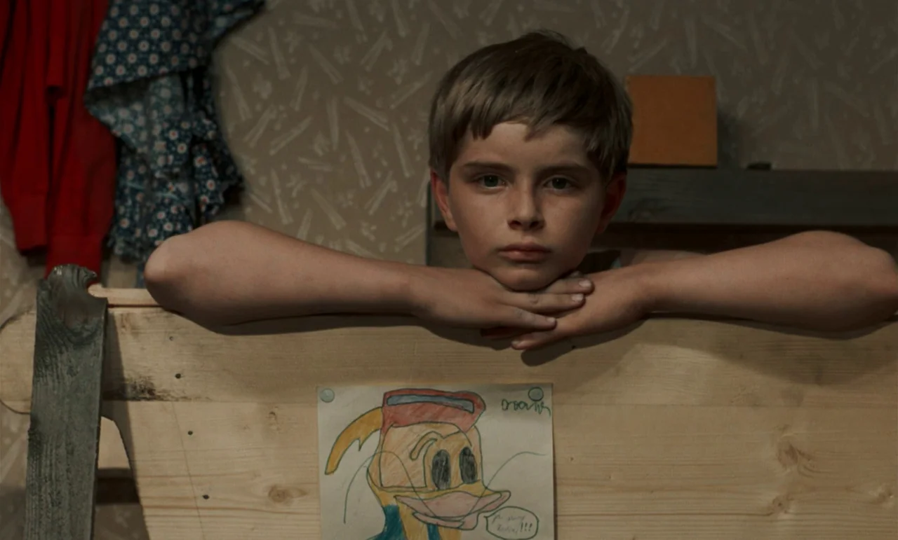 Larks On A String (1969) movie still: medium — A child sits behind a light wooden structure with patterned wallpaper on the wall. A hand-drawn image…