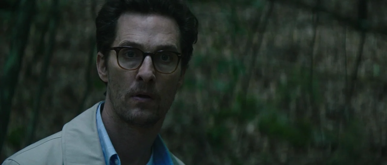 The Sea of Trees (2015) movie still: closeup — A man stands amidst dense, shadowed trees in a wooded area. He turns his head while wearing glasses…