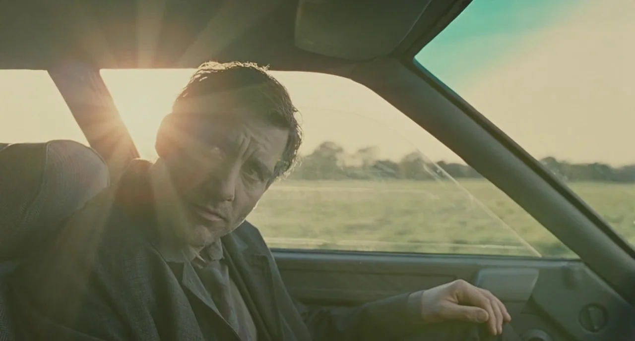 Children Of Men (2006) movie still: medium — A man sits inside the interior of a moving car during golden hour. Sunlight flares through the car…