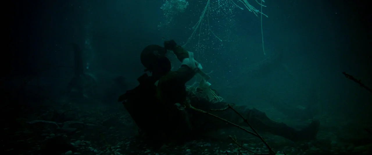 Overlord (2018) movie still: fullbody — A soldier lies on the rocky bed of a murky, dark green river. He is entangled in white ropes and reaches…