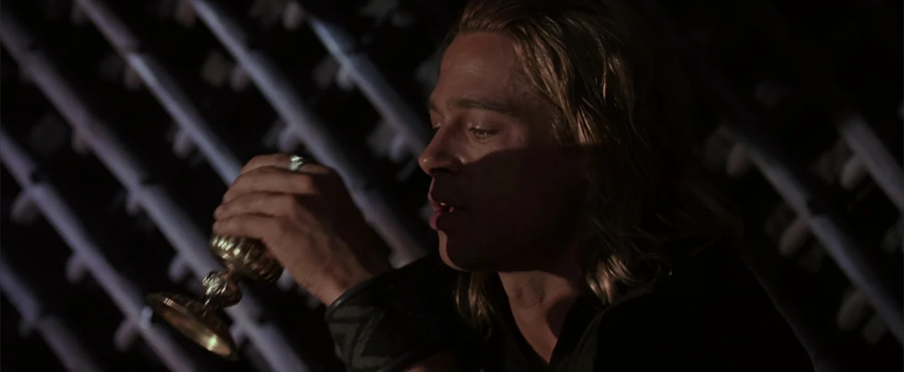 Troy (2004) movie still: closeup — A dark stone chamber features patterned walls in the background. A blonde man holds a golden goblet near his face.
