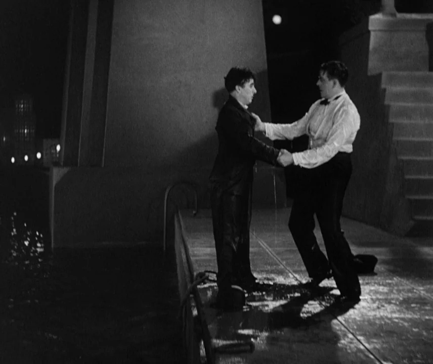 City Lights (1931) movie still: fullbody — A wet concrete pier at night with a large stone structure rising behind the men. A man in a dark suit…