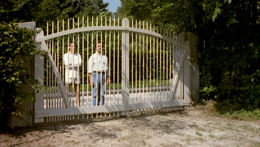 Funny Games (1997) movie still: fullbody — A white wooden gate stands in front of a dense line of trees. Two men stand behind the fence waiting.