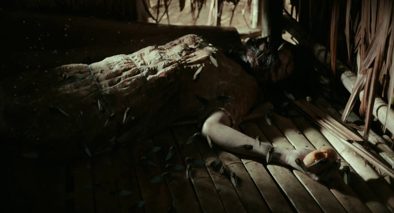 In My Mother’s Skin (2023) movie still: medium — A dark hut interior with a slatted bamboo floor and woven thatch walls. A woman lies prone on the…