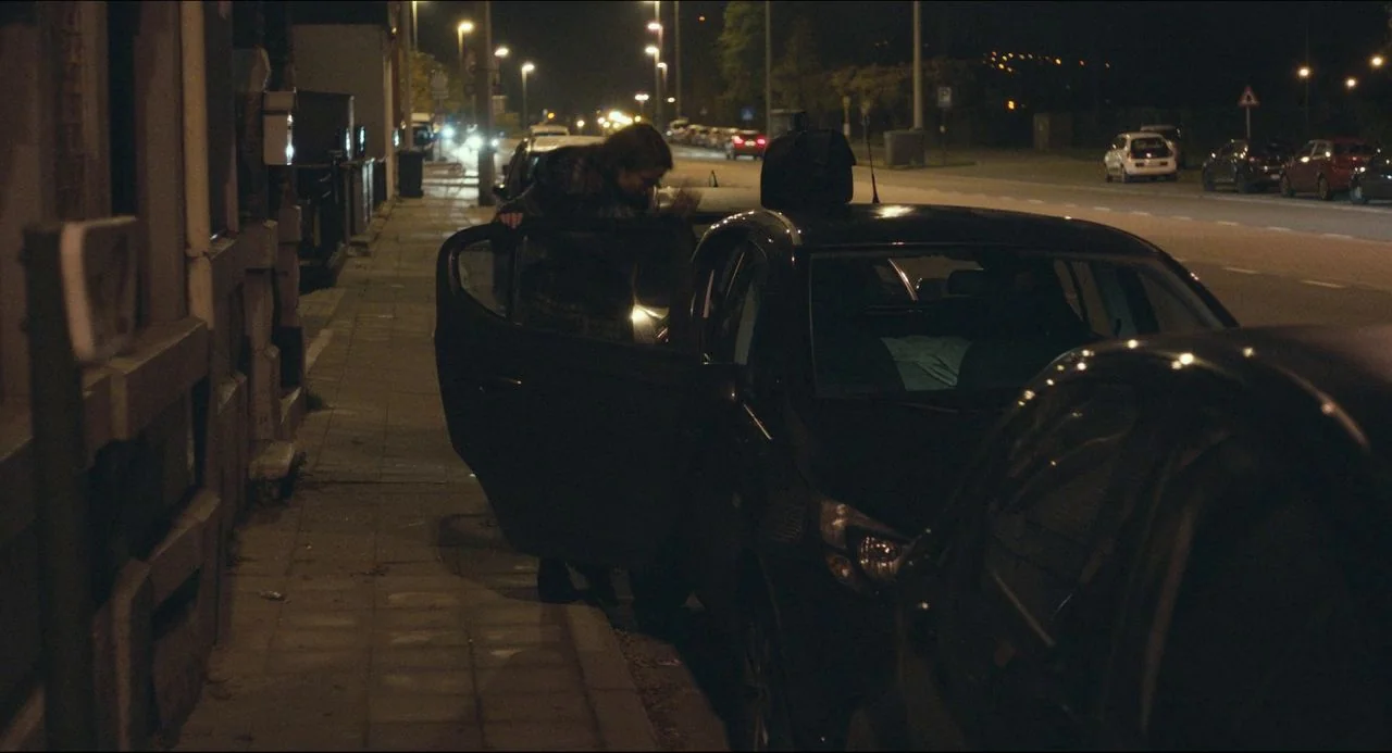 The Unknown Girl (2016) movie still: wide — A quiet urban roadside at night with parked vehicles along a narrow sidewalk. A woman bends to enter the…